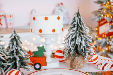 A charming table arrangement showcases a white lamp with orange polka dots, surrounded by small Christmas trees and festive ornaments, creating a cozy holiday atmosphere. clipart