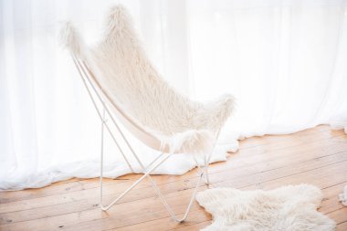 A butterfly chair featuring a plush white fur cover stands elegantly in a sunlit room with soft wooden floors and sheer curtains, adding a touch of comfort to the minimalist decor. clipart