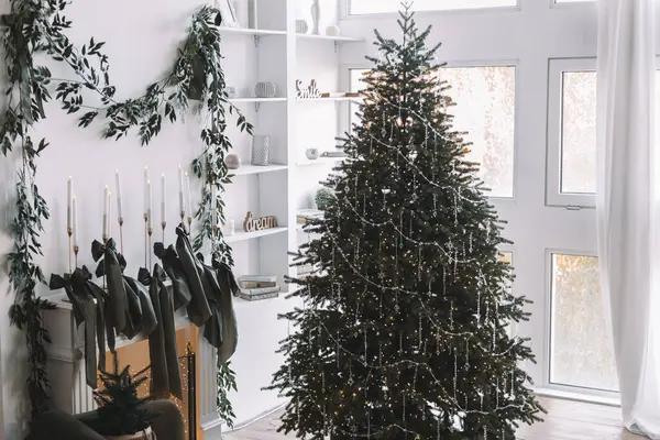 Güzel dekore edilmiş bir Noel ağacı, ışık ve süslemelerle süslenmiş rahat bir yaşam alanında duruyor. Çevreleyen yeşillik, odanın tatil cazibesini artırıyor..