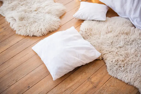 A cozy interior features soft white pillows scattered on a wooden floor, accompanied by fluffy white rugs, creating a warm and inviting atmosphere for relaxation.