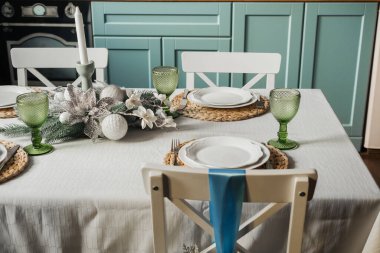 Güzel düzenlenmiş bir yemek masasında yeşil bardaklar, beyaz tabaklar ve içinde mumlar ve süsler olan zevkli bir süs eşyası bulunur..