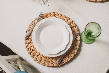 Çok güzel düzenlenmiş bir masa, yeşil cam ve paslanmaz çelik çatal bıçak eşliğinde doğal dokuma bir tabak üzerine kurulmuş, sakin bir yemek atmosferi yaratıyor..