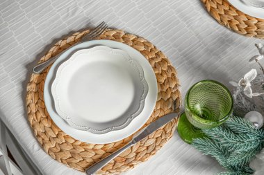 Güzelce düzenlenmiş bir yemek masasında örülmüş beyaz bir tabak bulunur. Yanında, yumuşak ışıkta parlayan yeşil bir cam yemek için sıcak ve davetkar bir atmosfer yaratır..