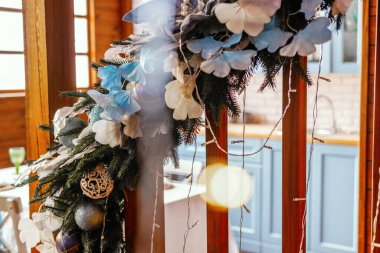 Güzel işlenmiş bir kış çelengi, beyaz çiçekler, mavi aksanlar ve parıldayan ışıklarla süslenmiş ahşap bir duvara asılarak şık bir mutfak ortamında rahat bir atmosfer yaratıyor..