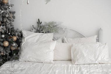 Huzurlu bir kış yatak odasında birden fazla yastığı olan yumuşak beyaz bir yatak sergilenir. Güzel süslenmiş bir Noel ağacı şenlikli bir dokunuş ekler ve huzurlu atmosferi güçlendirir..