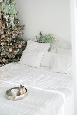 Konforlu bir yatak odası, beyaz çarşaflı ve gümüş tepsili, güzelce döşenmiş bir tatil ağacının yanında dinlenmek için mükemmel bir kış atmosferi yaratıyor..