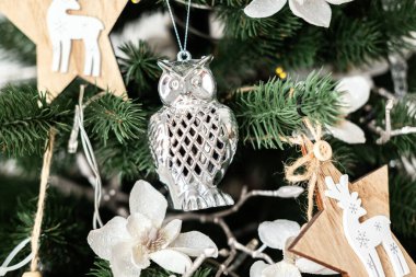 Tahta yıldızlar ve narin beyaz çiçeklerle çevrili yeşil bir Noel ağacına asılı gümüş bir baykuş süsü. Şenlikli dekorasyonlar neşeli bir tatil ortamı yaratıyor..