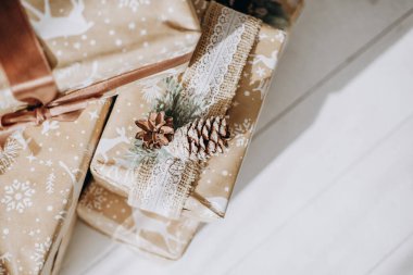 Dikkatlice paketlenmiş hediyeler kozalaklarla süslenmiş ve dantelli parlak ahşap bir yüzey üzerinde oturuyor. Şenlikli ambalaj, bayram sezonunun neşesini yakalıyor, kutlamaya ve hediye vermeye davet ediyor..
