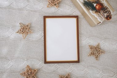 Dekoratif yıldızlar ve paketlenmiş bir hediyeyle çevrili boş bir çerçeve örülmüş bir masa örtüsüne yerleştirilir. Yumuşak dokular, tatil kutlamaları için uygun sıcak ve şenlikli bir ortam yaratıyor..