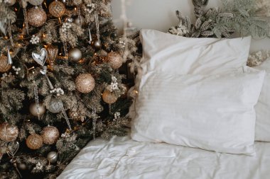 A cozy bedroom features a beautifully decorated Christmas tree next to a neatly made bed, adorned with soft white pillows, creating a warm holiday atmosphere. clipart