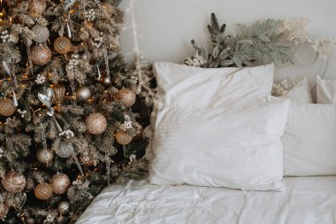 Sıcak bir kış yatak odası, güzel dekore edilmiş bir Noel ağacının yanında yumuşak beyaz yastıklarla süslenmiş bir yatak içerir. Neşeli atmosfer davetkar ve sıcak..
