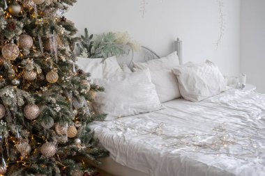 Sıcak ve davetkar bir yatak odasında güzel dekore edilmiş bir Noel ağacı ve yumuşak beyaz çarşaflarla süslenmiş bir yatak var. Bayramlar için mükemmel bir ortam yaratıyor..