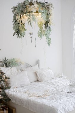 A peaceful bedroom features a neatly made bed with bright linens and hanging greenery overhead. Soft lights add a warm ambiance that complements the festive decorations in winter. clipart