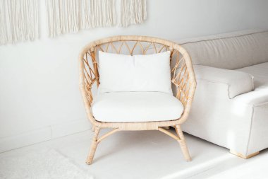 Pofuduk beyaz yastıklı şık bir rattan sandalye parlak bir oturma odasında oturur, açık renkli bir kanepe ve uzayın sıcaklığını ve rahatlığını arttıran minimalist bir dekorla tamamlanır..