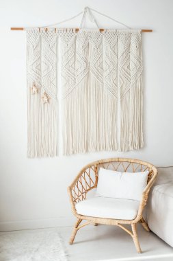 Çok güzel yapılmış bir makrom duvar, minimalist bir oturma odasını süslüyor, yanında şık bir rattan sandalye ve konfor ve sükunet öneren yumuşak dokular var.