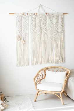 Çok güzel yapılmış bir makrom duvar, minimalist bir oturma odasını süslüyor, yanında şık bir rattan sandalye ve konfor ve sükunet öneren yumuşak dokular var.