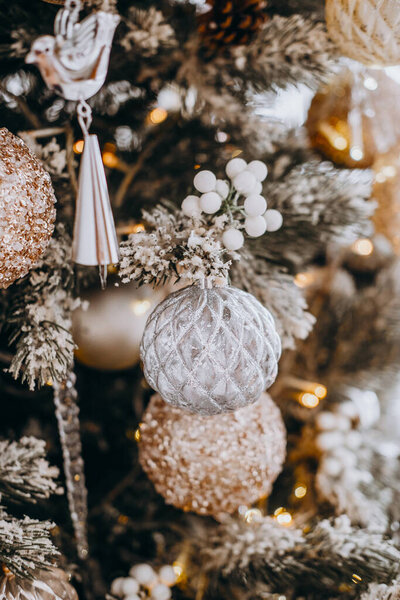 Beautifully crafted ornaments hang from a Christmas tree, featuring silver and gold tones. The soft glow of lights enhances the festive atmosphere of the holiday season.