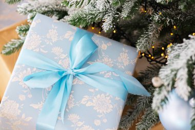Zarif bir şekilde paketlenmiş, açık mavi çiçekli bir hediye ışıldayan ışıklar ve süslemelerle süslenmiş gür, karlı bir Noel ağacının altında duruyor..