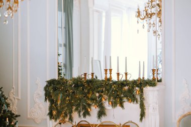 A beautifully arranged mantel adorned with evergreen garlands, twinkling lights, and elegant candlesticks creates a festive atmosphere in a light-filled room during the holiday season. clipart