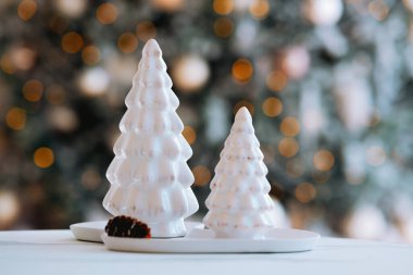 Two ceramic Christmas trees are displayed on a table, surrounded by a warm, festive background of twinkling holiday lights, creating a cozy seasonal atmosphere. clipart