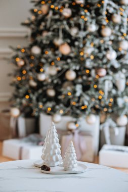 Delicate white ceramic trees are placed on a table in a warm room, framed by a beautifully decorated Christmas tree adorned with lights and ornaments, creating a festive atmosphere. clipart