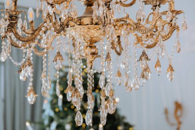 Karmaşık ayrıntıları ve parlak kristalleri olan çarpıcı bir avize zarifçe asılı duruyor, arka planda güzel dekore edilmiş bir tatil ağacının üzerine sıcak bir ışık tutuyor..