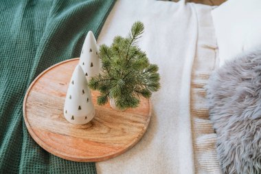 A round wooden tray holds two white ceramic Christmas trees and a small pine plant. Soft textiles in green and beige create a warm, inviting atmosphere in a cozy interior. clipart