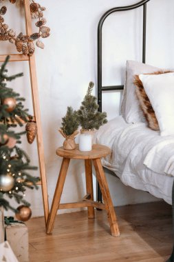 A charming bathroom corner showcases a wooden stool topped with a small evergreen arrangement beside a cozy bed adorned with soft pillows and decorative accents for winter. clipart