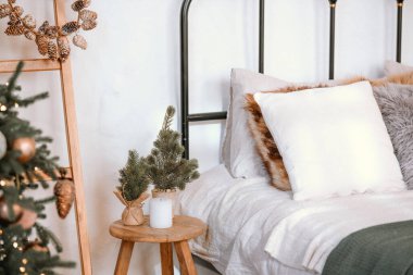 Sıcak ve davetkar bir yatak odasında yumuşak yastıklarla süslenmiş bir yatak ve ahşap bir taburede küçük bir ağaç bulunur. Tatil dekoru rahatlatıcı alana cazibe katıyor, rahat bir ortam yaratıyor..