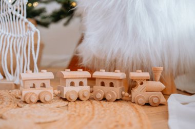 A charming wooden toy train made of natural materials sits on a soft rug, with a festive Christmas tree in the background, creating a warm and inviting holiday atmosphere. clipart