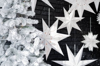 Beyaz kağıt yıldızlar karanlık bir duvardan zarif bir şekilde sarkarak buzlu bir Noel ağacının yanında şenlikli bir atmosfer yaratırlar. Dekorasyon, tatil sezonu için mükemmel bir kış cazibesi yayıyor..
