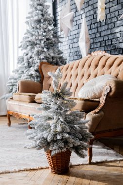 Konforlu bir oturma odasında eski bej bir kanepenin yanındaki sepette küçük bir buzlu Noel ağacı bulunur. Mekan, doğal ışığın içeri aktığı şenlikli bir şekilde dekore edilmiş..