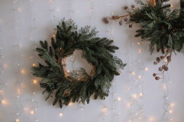 Sonsuza dek yeşil dallardan ve çam kozalaklarından oluşan iki çelenk bir duvarı süslüyor. Yumuşak parıldayan ışıklarla aydınlatılıyor. Kış kutlamaları için mükemmel bir tatil ortamı yaratıyor..