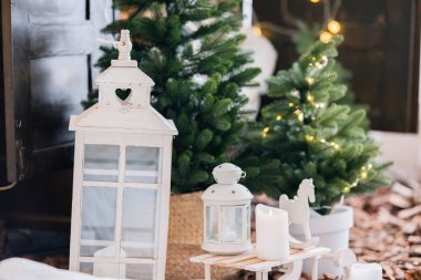 Kapalı bir kış dekorasyonunda beyaz fenerler, mumlar ve parıldayan ışıkları olan küçük Noel ağaçları, tatil sezonu için sıcak ve şenlikli bir atmosfer yaratıyor..