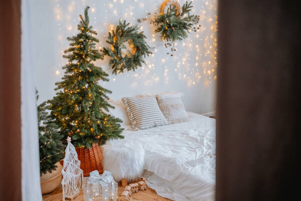 A warm and inviting bedroom features small Christmas trees, twinkling lights, and festive wreaths on the wall, creating a joyful holiday atmosphere perfect for relaxation.