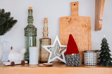 A charming display of holiday decorations on a wooden shelf features glass bottles, festive figurines, and a wooden cutting board. The atmosphere is warm and inviting, perfect for the season. clipart