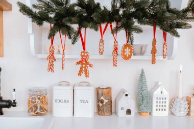 Samimi bir mutfakta, yukarıdan sarkan zencefilli kurabiyelerle dolu bir Noel ruhu sergileniyor. Aşağıda, şeker ve çay kavanozları büyüleyici dekoratif evlerin yanında oturur..