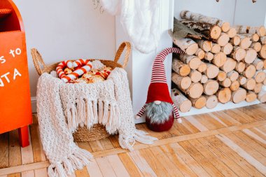 A festive corner showcases a gnome in a red hat next to a basket filled with striped blankets. The wooden logs in the background add to the cozy winter ambiance of the room. clipart