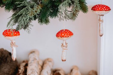 Three decorative mushroom ornaments with red caps and white spots dangle from green pine branches in a cozy indoor setting, adding a whimsical touch to the decor. clipart