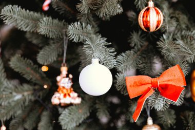 Güzel dekore edilmiş bir Noel ağacı, beyaz bir küre ve parlak kırmızı bir fiyonk da dahil olmak üzere çeşitli süsler sergiler. Atmosfer şenlikli, tatil sezonunun ruhunu yansıtıyor..