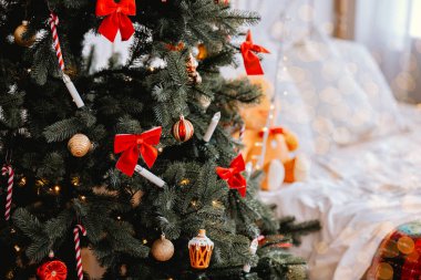 Güzel süslenmiş bir Noel ağacı rahat bir yatak odasında duruyor, kırmızı kurdeleler ve çeşitli süsler sergiliyor. Yumuşak beyaz yatak ve sıcak ışık şenlikli bir atmosfer yaratır..