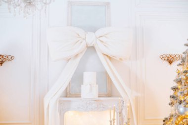 Noel için süslenmiş rahat bir odada bir şöminenin üzerinde büyük dekoratif bir yay ve yanında da parlak bir ağaç bulunuyor. Ortam, tatil sezonunun neşesini mükemmel bir şekilde yakalıyor..