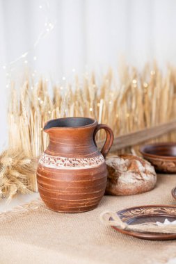 Kaba bir çömlek sürahisi ve dekoratif tabaklar desenli bir yüzeye yerleştirilir. Kurutulmuş buğday sapları sıcak bir dokunuş ekliyor. Sıcak bir kış atmosferi yaratıyor. Tatil ve toplanma için mükemmel..