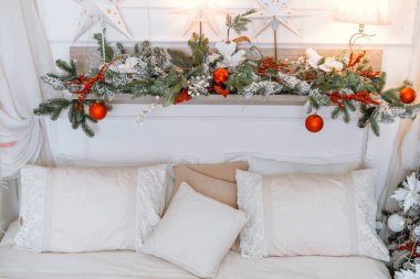 Sıcak bir kış ayarı, yumuşak yastıklı, düzgünce yapılmış bir yatağın üzerinde kırmızı süslemeler ve beyaz çiçeklerle süslenmiş şenlikli bir Noel çelengi içerir. Dekor sıcak bir tatil ortamı yaratıyor..