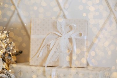 A lovely gift box wrapped in white with a delicate bow sits near a Christmas tree. Soft winter lights twinkle, creating a festive ambiance for Christmas and New Year celebrations. clipart