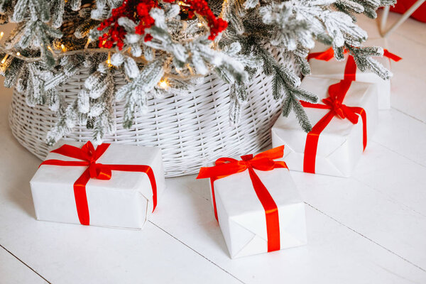 Ярко-белые подарочные коробки с красными лентами помещаются под снежной елкой. Настройка излучает теплую зимнюю атмосферу, празднуя радость Рождества и Нового года.