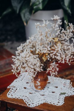 Kurumuş çiçeklerle dolu kırsal bir vazo ahşap bir masanın üzerindeki dantel bir altlığın üzerinde duruyor. Dekor, sıcaklığı ve nostaljiyi davet eden doğal dokuları ve toprak tonlarını gösteren rahat bir atmosfer yaratıyor..