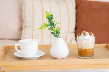 Krem şanti ve yeşil bitkiyle kaplı cam bir tatlının yanında beyaz bir fincan oturuyor..