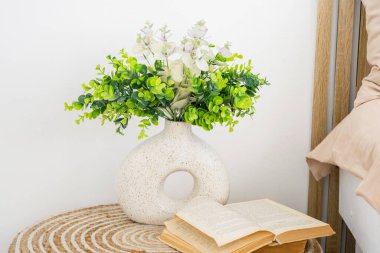 A stylish vase filled with greenery sits next to an open book on a nightstand. clipart