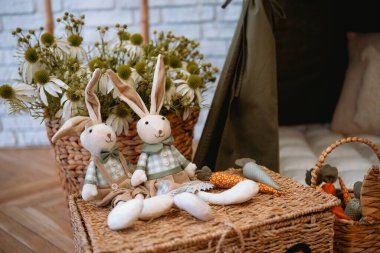 İki oyuncak bebek, süslü havuç ve çiçeklerle çevrili dokuma bir sepetin üzerinde otururlar..